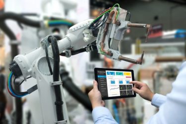 Hand eines Ingenieurs bedient ein Tablet, um in Echtzeit einen schweren Roboterarm zu steuern