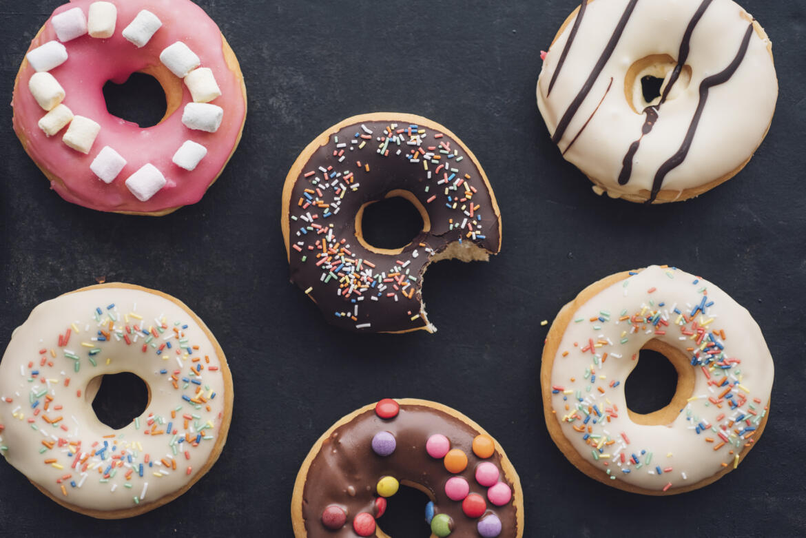 Sechs bunt-verzierte Donuts vor einem schwarzen Hintergrund. Der Donut in der Mitte ist angebissen.