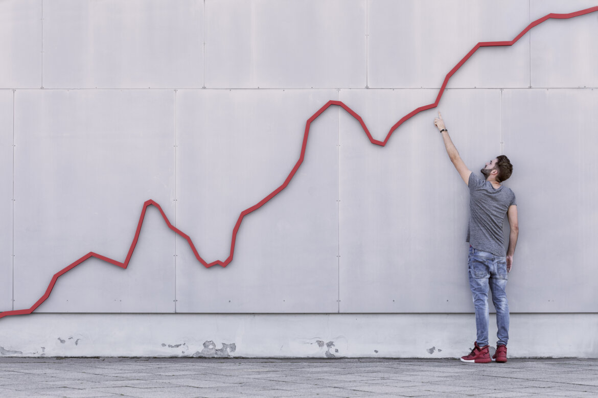 A red bar line graph rises like a financial graph or graphic with a young man pointing at the growth against a wall, Foto: © Jonathan Kitchen / Getty