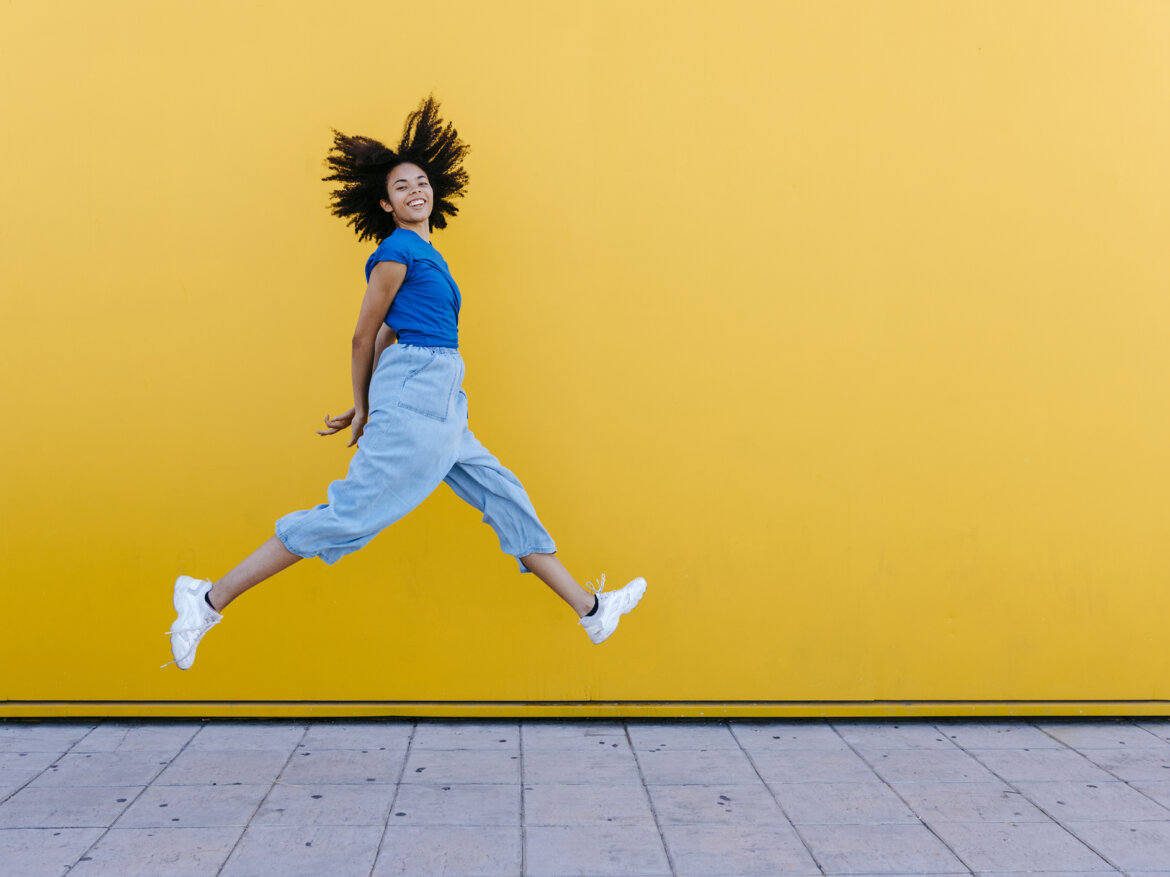 Junge afroamerikanische Frau in blauer Kleidung springt vor gelber Wand