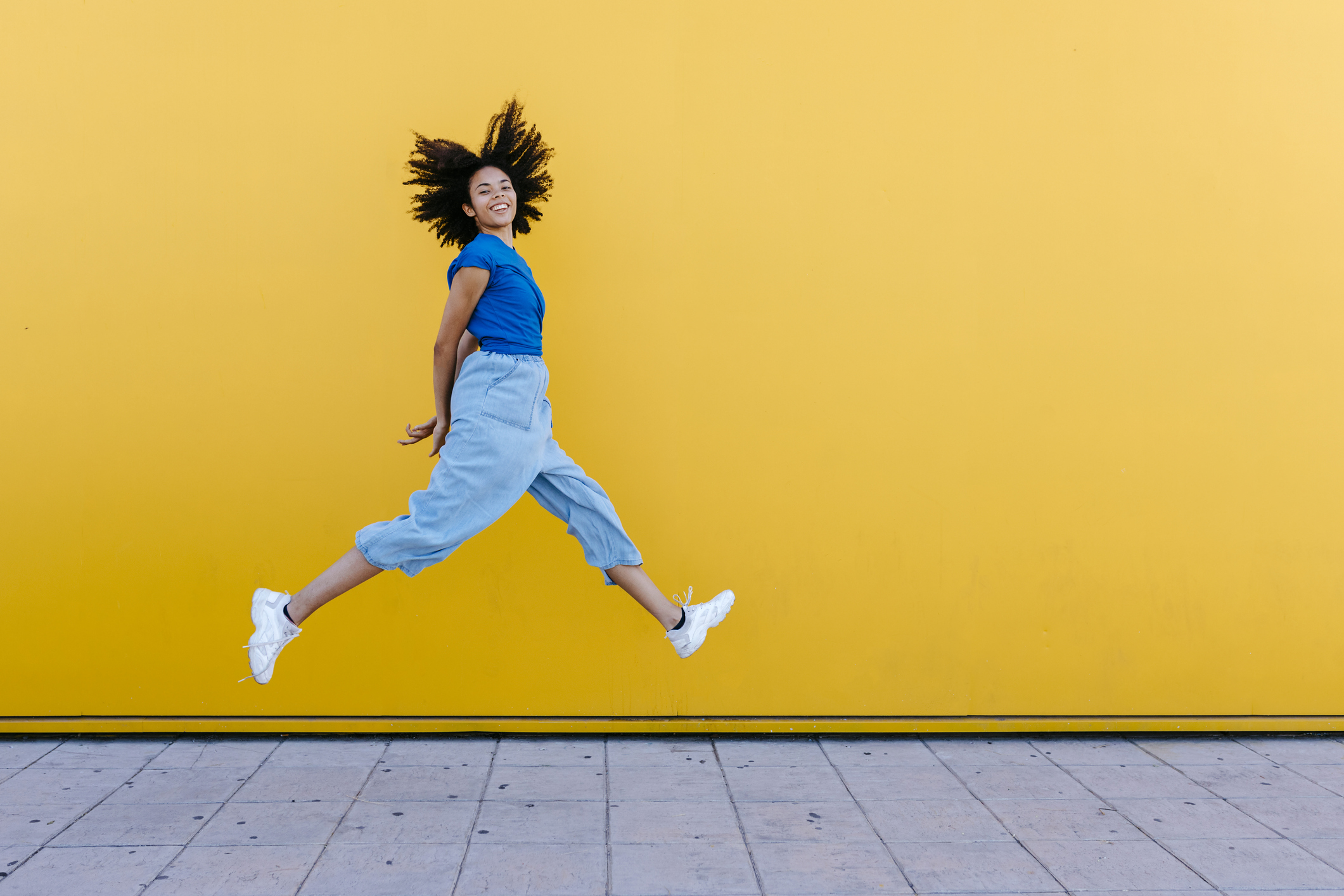 Junge afroamerikanische Frau in blauer Kleidung springt vor gelber Wand